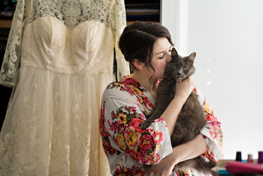 A bride kisses her cat in front of her wedding dress.